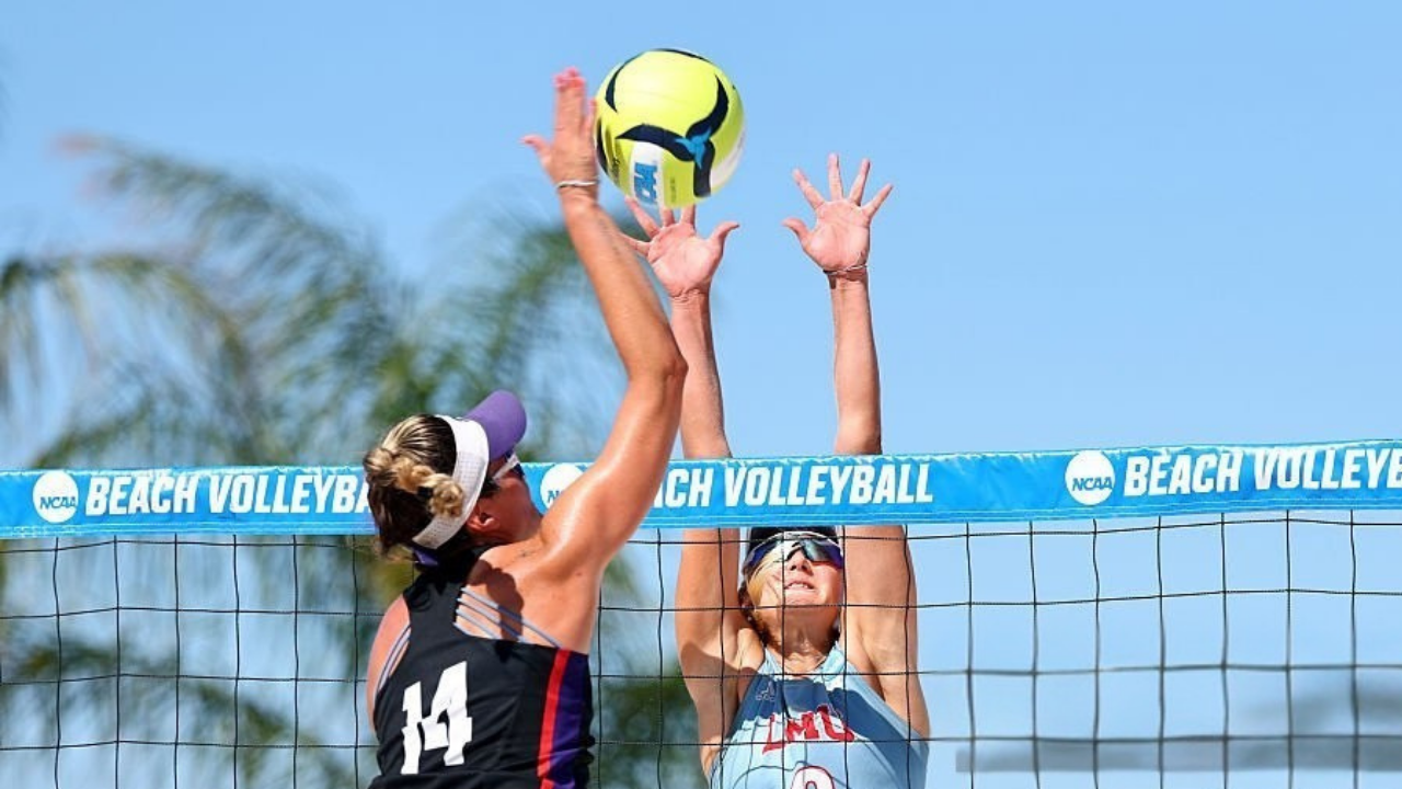 "An elite D1 beach volleyball player in a black jersey (#14) rises high above the net to block an attack during an NCAA championship match, showcasing the peak athletic skill required at the D1 level." Get Help fromSalava Volleyball.